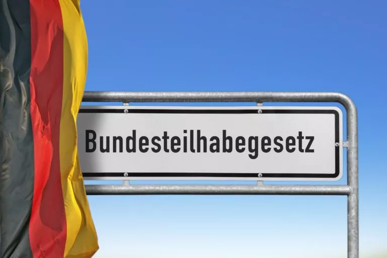 Auf diesem Bild sehen Sie einen Ausschnitt aus dem E-Learning-Kurs zum Thema Bundesteilhabegesetz. Auf dem Bild sehen Sie ein Straßenschild, auf dem Bundesteilhabegesetz steht. Daneben die Deutschlandflagge.