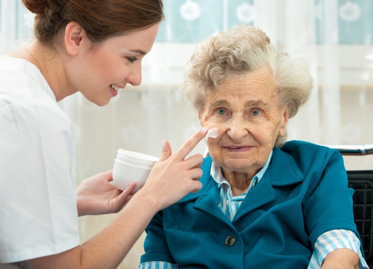Auf diesem Bild sehen Sie einen Ausschnitt aus dem E-Learning-Kurs zum Thema Hygiene in der ambulanten Pflege. Auf dem Bild ist eine ältere Frau zu sehen, der von einer Pflegerin das Gesicht eingecremt wird.
