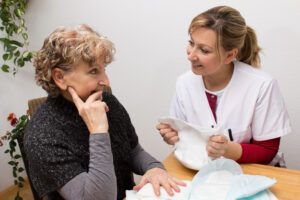 Auf diesem Titelbild des Blogartikels zum Thema Expertenstandard Kontinenzförderung in der Pflege sehen Sie eine Pflegerin die sich mit einer älteren Frau unterhält und dabei eine Inkontinenzeinlage in ihren Händen hält.