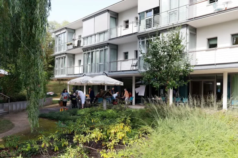 Pflegeeinrichtung mit Terrasse, Sonnenschirmen und Bewohnern im Grünen.