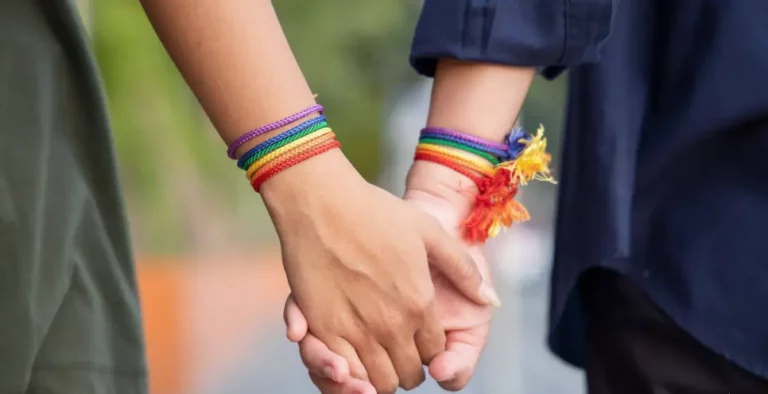2 Personen halten Hände, beide tragen regenbogenfarbige Armbänder.