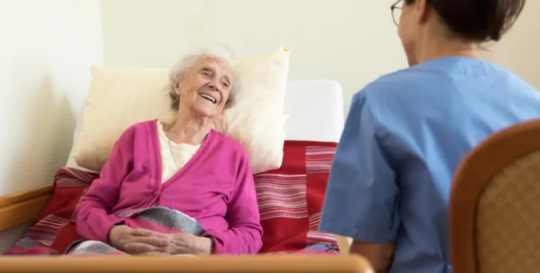 Eine Pflegeperson sitzt vor einem Bett mit älterer Dame darin und unterhält sich mit ihr freudig.