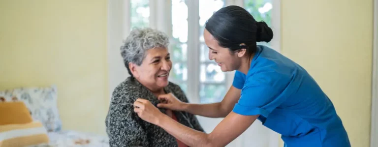 Pflegeperson hilft sitzender Person beim Anziehen einer Strickjacke.