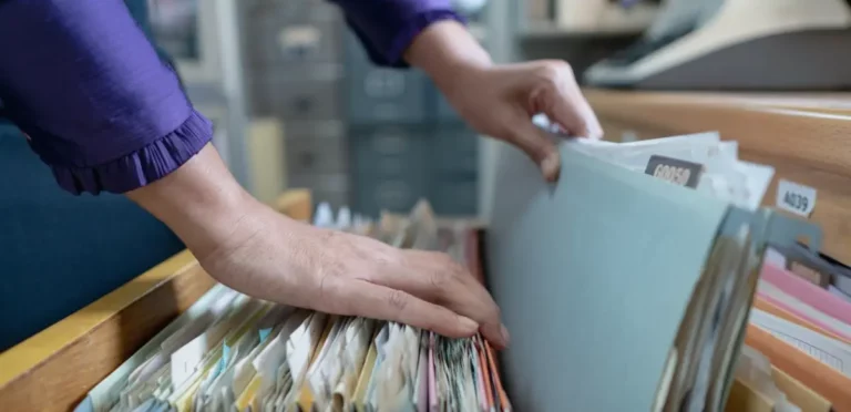 Hände blättern durch Hängeregister mit Akten in einer Holzschublade.