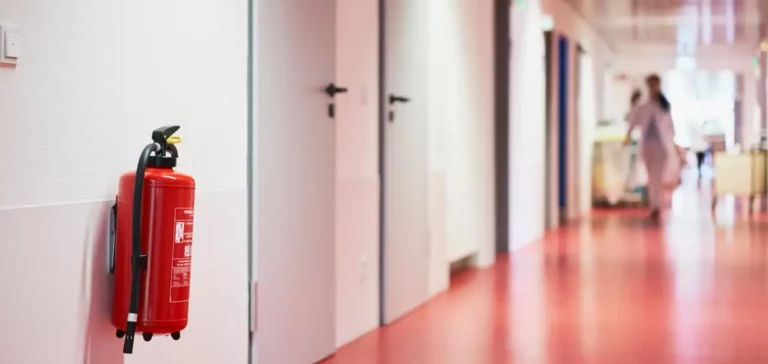 Roter Feuerlöscher im Krankenhausflur mit unscharfer Person im Hintergrund.