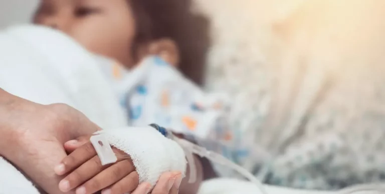 Kind liegt im Pflegebett mit Infusion am Arm und wird an der Hand gehalten.