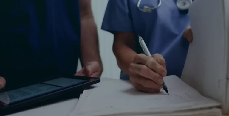 Pflegekraft dokumentiert Patientendaten schriftlich auf Papier neben einer digitalen Patientenakte auf einem Tablet.