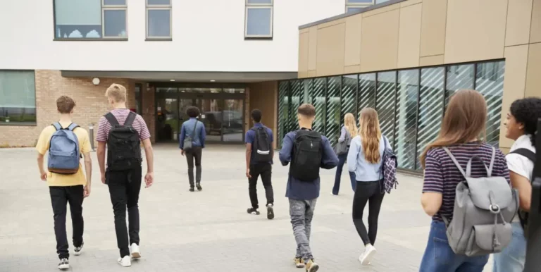 Gruppe Jugendlicher mit Rucksäcken geht auf Schulgebäude mit Glastüren zu.