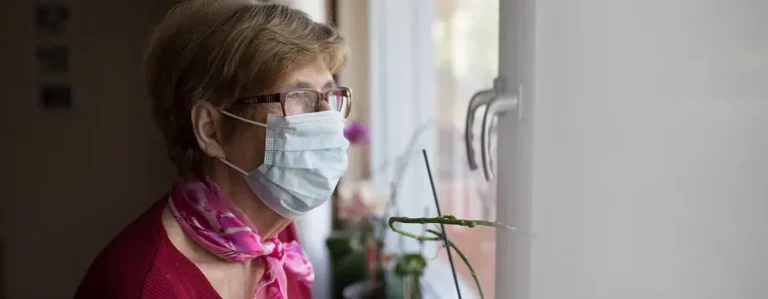 Ältere Frau mit OP-Maske schaut nachdenklich aus dem Fenster
