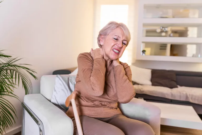 Seniorin sitzt mit schmerzverzerrtem Gesicht auf einem Sessel und hält sich den Nacken.