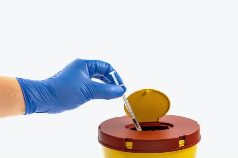 Hand mit blauen Handschuhen entsorgt Spritze in Sicherheitsbehälter.