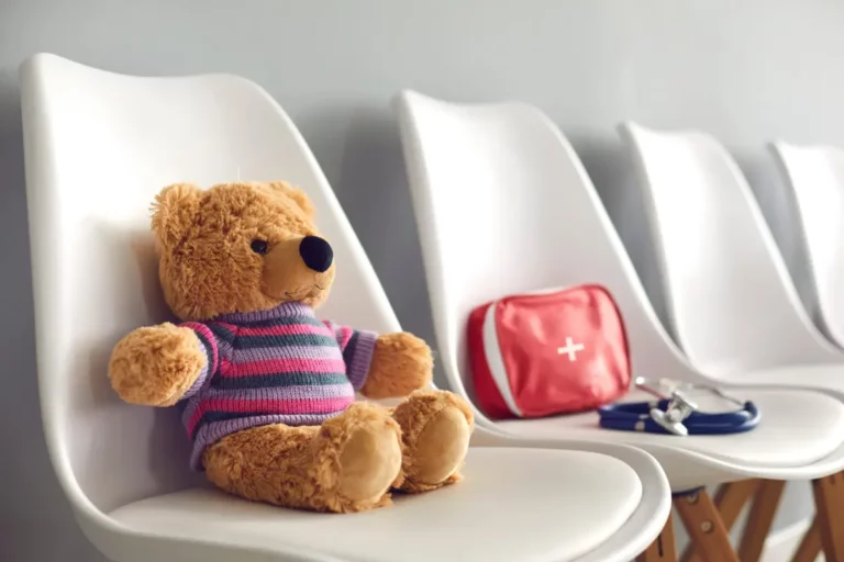 Teddybär sitzt auf Stuhl neben Erste-Hilfe-Set im Wartezimmer.