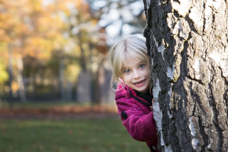Auf dem Bild ist ein kleines, blondes Mädchen zu sehen, das sich hinter einem Baum versteckt und lacht. Sie trägt eine lila Kapuzenjacke.