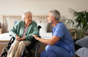 Pflegeperson kniet neben einer älteren Dame im Rollstuhl. Sie unterhalten sich.