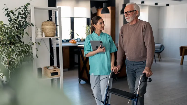 Pflegeperson steht neben älterer Person mit Rollator im Wohnraum.