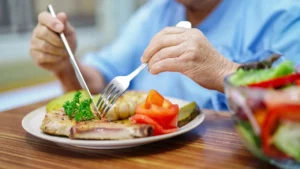Hände einer älteren Person halten Messer und Gabel über einem Teller mit Essen.