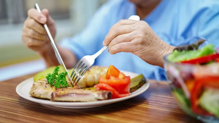 Hände einer älteren Person halten Messer und Gabel über einem Teller mit Essen.