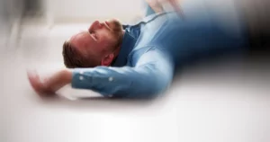 Person liegt am Boden in Innenraum, Augen geschlossen, Oberkörper sichtbar, Bild verschwommen