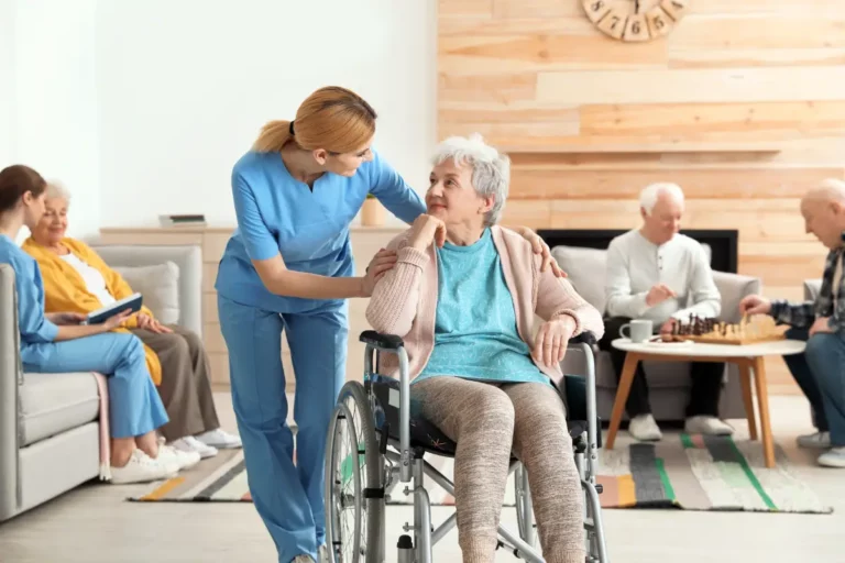 Pflegeperson betreut Person im Rollstuhl in Gemeinschaftsraum mit älteren Menschen.